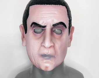 Può includere: Maschera integrale in lattice raffigurante il volto di un uomo con capelli e sopracciglia scure. La maschera ha un incarnato pallido, occhi grigi e una bocca leggermente contratta. La maschera è progettata per essere indossata.