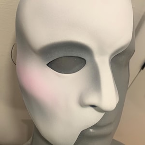 Può includere: Una maschera teatrale, dipinta in bianco e grigio, poggia su una testa di manichino. La maschera copre metà del viso, con un naso prominente e un'apertura per l'occhio. Un delicato fard rosa accentua la guancia. Il design generale suggerisce il tema del Fantasma dell'Opera.