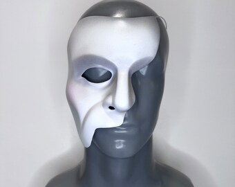May include: A white half mask with a gray mannequin head. The mask covers the right side of the face and has a long, pointed nose.