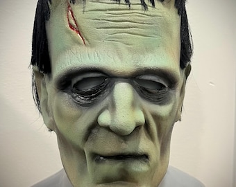 May include: A green and gray Frankenstein monster mask with black hair and a scar. The mask has a realistic design with a prominent brow, sunken eyes, and a stitched-up neck. A Halloween costume accessory.