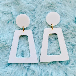 May include: A pair of white geometric earrings with a gold hoop. The earrings are shaped like a trapezoid with a rectangular cutout in the center.
