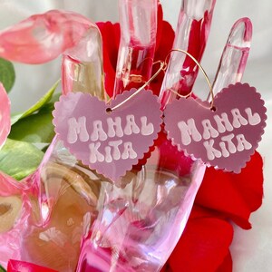 Op de afbeelding: Een paar roze hartvormige oorbellen met gouden hoepels. De harten hebben de tekst "Mahal Kita" in wit.