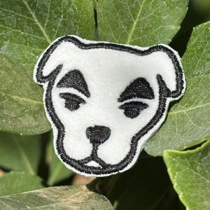 以下が含まれることがあります： 黒い目、鼻、輪郭などの黒いアクセントが付いた白い犬の顔のパッチ。パッチは緑の葉に囲まれています。衣類やアクセサリーに加えるのに最適です。