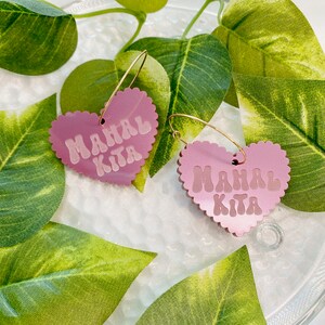 Op de afbeelding: Een paar roze hartvormige creolen met de tekst "Mahal Kita" in witte letters. De oorbellen liggen op een wit oppervlak met groene bladeren.