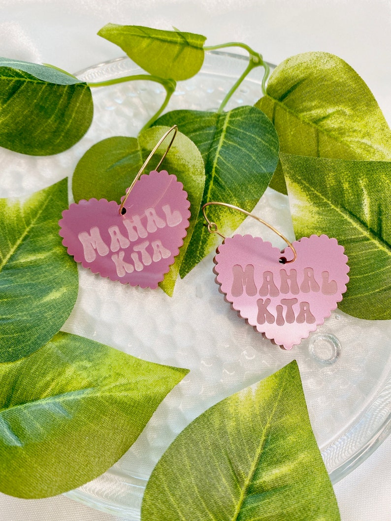 Op de afbeelding: Een paar roze hartvormige creolen met de tekst "Mahal Kita" in witte letters. De oorbellen liggen op een wit oppervlak met groene bladeren.