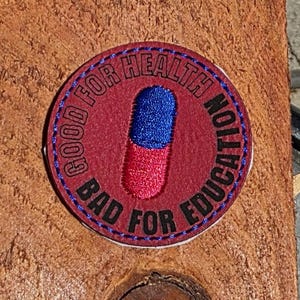 May include: A circular patch with the text "GOOD FOR HEALTH, BAD FOR EDUCATION" encircling a red and blue pill design. The patch is red with blue and white stitching.