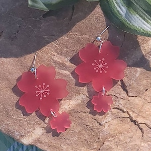 May include: A pair of dangle earrings featuring two pink flower charms. The larger flower charm has a white centre with a five-pointed star design. The smaller flower charm is a solid pink.