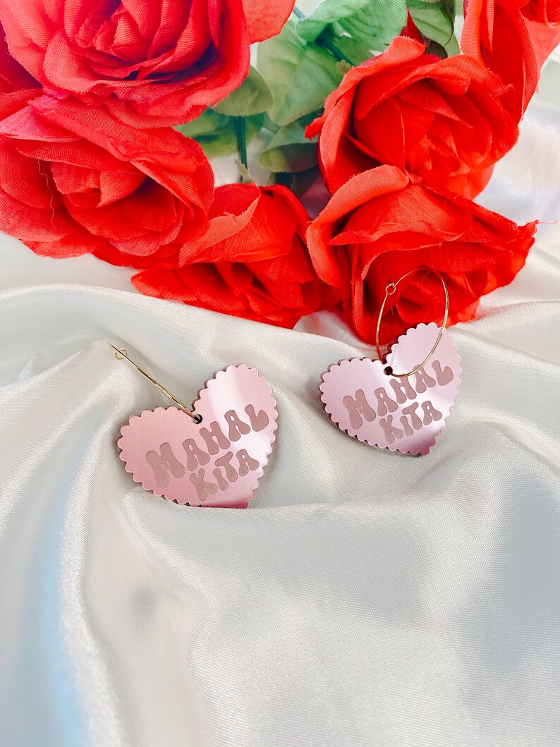 Op de afbeelding: Een paar roze hartvormige oorbellen met de tekst "Mahal Kita" in een retro-lettertype. De oorbellen hangen aan gouden hoepels.
