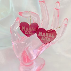 Op de afbeelding: Een paar roze hartvormige oorbellen met de tekst "Mahal Kita" in witte letters. De oorbellen staan op een roze displaystandaard.