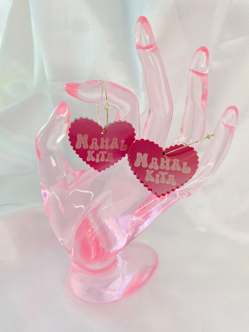 Op de afbeelding: Een paar roze hartvormige oorbellen met de tekst "Mahal Kita" in witte letters. De oorbellen staan op een roze displaystandaard.