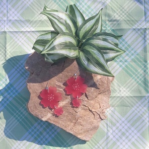 May include: A pair of red cherry blossom earrings with a smaller flower charm hanging below the larger flower. The earrings are on a rock with a green plant in the background.
