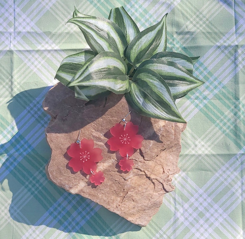 May include: A pair of red cherry blossom earrings with a smaller flower charm hanging below the larger flower. The earrings are on a rock with a green plant in the background.