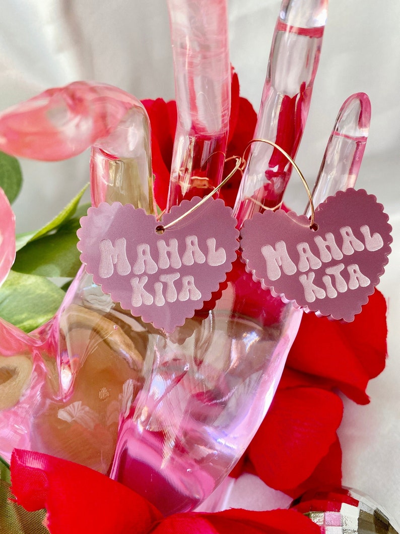 Op de afbeelding: Een paar roze hartvormige oorbellen met gouden hoepels. De harten hebben de tekst "Mahal Kita" in wit.