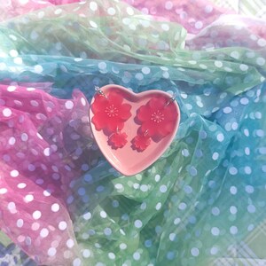 May include: A pair of pink cherry blossom earrings displayed on a pink heart-shaped dish. The earrings are made of resin and feature a delicate floral design.