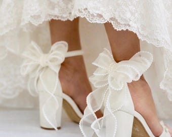 Può includere: Un paio di sandali color avorio con tacco largo e un ampio cinturino alla caviglia decorato con un grande fiocco in organza trasparente. I bordi del fiocco sono rifiniti con delicate perle bianche, indossati sotto un abito in pizzo bianco.