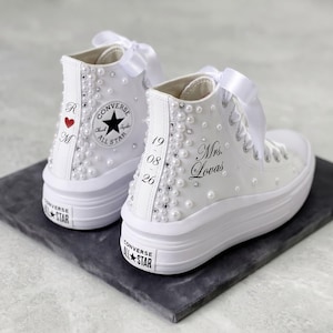 Könnte beinhalten: Weiße High-Top-Plateau-Sneaker, verziert mit Kunstperlen und Strasssteinen. Sie verfügen über einen weißen Schnürverschluss mit Schleife. Auf den Sneakern steht der Text "Mrs. Lovas" und das Datum "19 08 26". Das Converse All Star-Logo ist sichtbar.
