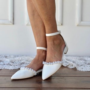 May include: White satin bridal shoes with a pointed toe and ankle strap. The shoes are decorated with a row of faux pearls around the edge of the toe.