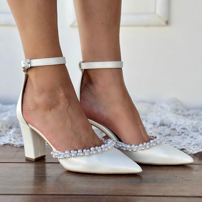 May include: White satin bridal heels with a pointed toe and a chunky block heel. The shoes are adorned with a row of faux pearls around the ankle strap and the toe.