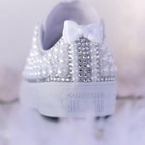 K&ouml;nnte beinhalten: Wei&szlig;e Canvas-Sneaker mit einer wei&szlig;en Schleife aus Satin und verziert mit Perlen und Strasssteinen.
