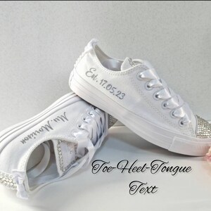 K&ouml;nnte beinhalten: Wei&szlig;e Canvas-Sneaker mit Strasssteinen an Zehen und Ferse. Die Sneaker sind mit dem Text "Est. 17.05.23" auf einem Schuh und "Mrs. Morrison" auf dem anderen personalisiert.