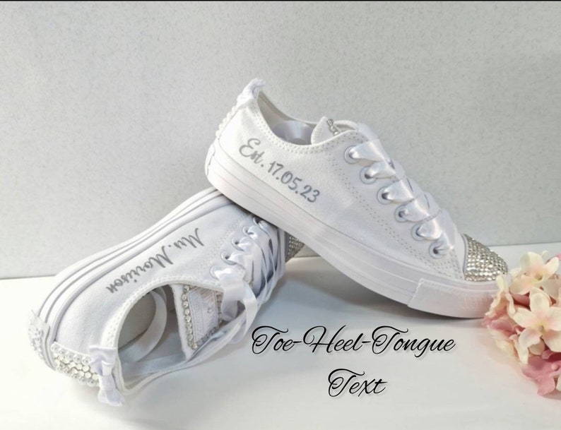 K&ouml;nnte beinhalten: Wei&szlig;e Canvas-Sneaker mit Strasssteinen an Zehen und Ferse. Die Sneaker sind mit dem Text "Est. 17.05.23" auf einem Schuh und "Mrs. Morrison" auf dem anderen personalisiert.