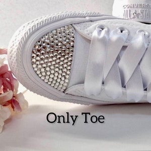 K&ouml;nnte beinhalten: Wei&szlig;e Canvas-Sneaker mit wei&szlig;en Satinband-Schn&uuml;rsenkeln. Die Zehenkappe des Schuhs ist mit klaren Strasssteinen besetzt. Der Text "Only Toe" ist unter dem Schuh aufgedruckt.
