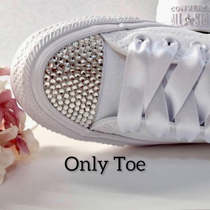 K&ouml;nnte beinhalten: Wei&szlig;e Canvas-Sneaker mit wei&szlig;en Satinband-Schn&uuml;rsenkeln. Die Zehenkappe des Schuhs ist mit klaren Strasssteinen besetzt. Der Text "Only Toe" ist unter dem Schuh aufgedruckt.