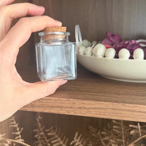 May include: A small, clear glass jar with a square shape and a cork stopper. The jar is held in a hand, with a wooden shelf and decorative items in the background. The jar is empty and ready to be filled.
