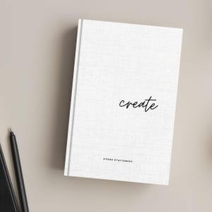 May include: White notebook with a gray spine and the word "create" written in black cursive. The bottom of the notebook says "Hygge Stationery".