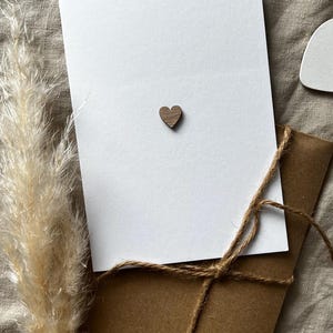 Puede incluir: Una tarjeta rectangular blanca con un pequeño corazón de madera marrón oscuro centrado. La tarjeta se coloca sobre un sobre de papel kraft marrón atado con cordel de yute fino, acompañado de una pluma de hierba de la pampa beige seca.
