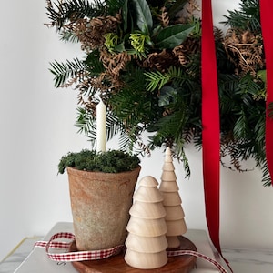 May include: A festive arrangement featuring a green Christmas wreath with red ribbon, a terracotta pot with a white candle, and two wooden Christmas trees. The scene is set on books titled "SCANDIRUSTIC" and "THE KINFOLK HOME".