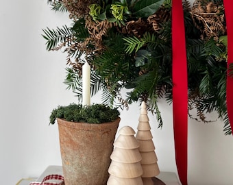 May include: A Christmas-themed still life featuring a green wreath with red ribbon, a terracotta pot with a white candle, and two wooden Christmas trees. The scene is set on books with the titles "SCANDIRUSTIC" and "THE KINFOLK HOME".