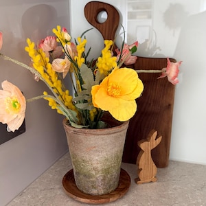 May include: A terracotta pot with a variety of colorful artificial flowers, including yellow poppies and peach-colored blooms. A wooden cutting board and a wooden bunny figurine are in the background. The arrangement sits on a light-colored countertop.