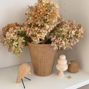Puede incluir: Una maceta de terracota llena de hortensias secas en tonos beige, rosa y verde. Aves decorativas de madera y una escultura de madera apilada se encuentran en una estantería blanca, creando una estética natural y rústica.