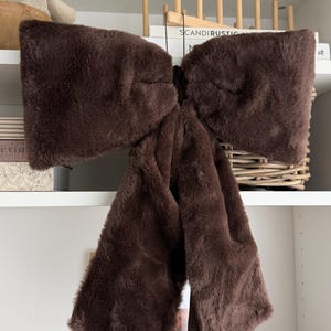 May include: A large, dark brown bow accessory crafted from soft, plush faux fur material. The bow features two wide, horizontal loops and two long, vertical tails that hang downwards, creating a voluminous and textured appearance.