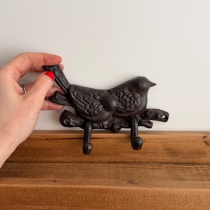 May include: A dark brown cast iron wall hook, shaped like a bird on a branch, with two hooks. The bird has detailed feather and eye features. The hook is mounted on a wooden shelf. The hook is designed for hanging items.