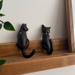 Könnte beinhalten: Zwei schwarze Katzenhaken auf einem Holzregal. Die Haken sind aus Gusseisen mit matter Oberfläche. Eine Katze sitzt, die andere blickt in die entgegengesetzte Richtung. Dekorativ und funktional.