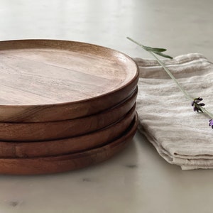 Puede incluir: Una pila de cuatro platos redondos de madera, con un acabado de veta de madera natural. Los platos están apilados sobre una superficie blanca, con una ramita de lavanda sobre una servilleta de lino beige a la derecha.