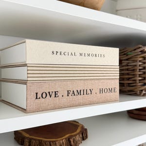 May include: Two decorative storage boxes with the text "SPECIAL MEMORIES" and "LOVE. FAMILY. HOME" stacked on a white shelf. The boxes have a neutral color scheme with a linen-like texture. A wooden bowl and a woven basket are also visible.