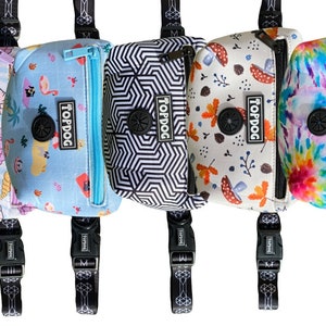 May include: Five different patterned dog waste bag dispensers with black straps and clips. The dispensers have a variety of patterns including seashells, geometric shapes, mushrooms, and tie-dye. Each dispenser has a black button with the word "Topdog" printed on it.