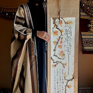 Op de afbeelding: Een bruin en beige gestreepte robe met een zwart onderhemd. De persoon houdt een rol met kalligrafie en een schilderij van een tak met oranje bladeren. De tekst op de rol luidt "الا الله عاله الا الله هو مولانا وَ عَليم فلينو الله تضمين القلوب  心の安らぎず得られないは アッラーを人念することにより を唱念し心の安らぎを得る。 これらの信仰したちはアッラー"