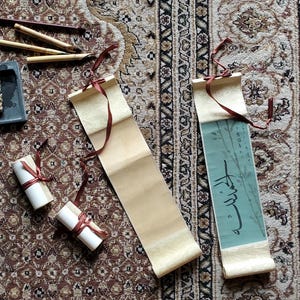 Könnte beinhalten: Draufsicht auf Kalligraphie-Werkzeuge und Schriftrollen auf einem gemusterten Teppich. Die Artikel umfassen Bambusstifte, einen grauen Tintenstein und gerollte Schriftrollen, die mit roten Bändern gebunden sind. Eine Schriftrolle hat türkisfarbenes Papier mit arabischer Schrift.