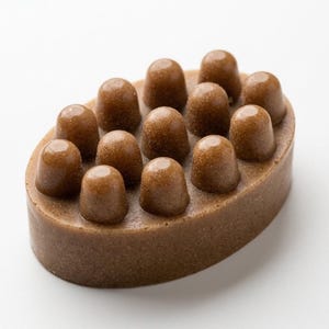 May include: An oval-shaped brown massage soap bar featuring a textured surface with twelve rounded, raised nubs arranged in a grid pattern. The soap has a speckled, earthy appearance and a smooth, solid base, designed for use as a handheld tool.