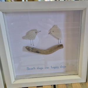 May include: A framed artwork featuring two stylized birds made of white material perched on a piece of driftwood. The artwork is set against a white background, with the phrase "Beach days are happy days" in blue script.