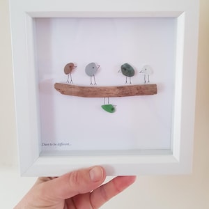 Pode incluir: Uma obra de arte emoldurada com pássaros de seixos em um galho de madeira flutuante. Os pássaros são de várias cores, incluindo marrom, cinza e verde. A obra está em uma moldura branca, e o texto "Dare to be different..." está na parte inferior.