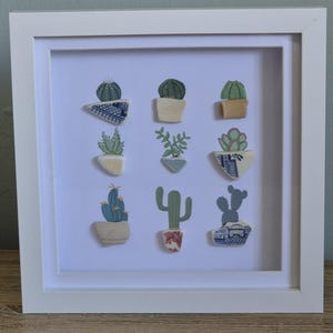 Op de afbeelding: Een ingelijst kunstwerk met negen kleine, handgemaakte cactus- en vetplantontwerpen. De planten staan in diverse potten en bakken, met groene, blauwe en beige tinten. De witte lijst omringt het kunstwerk, dat tegen een witte achtergrond is geplaatst.