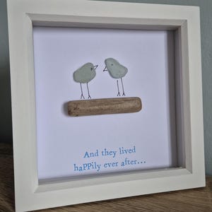 May include: A framed picture with two grey sea glass birds on a piece of driftwood. The white frame surrounds a white background with the blue text, "And they lived happily ever after..."