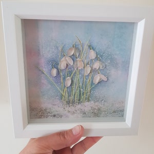May include: A framed artwork depicting snowdrop flowers in a watercolor style. The white flowers and green stems are set against a soft blue and purple backdrop. The artwork is presented in a white square frame.