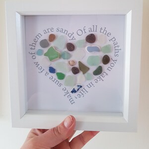 Puede incluir: Una obra de arte enmarcada que presenta una disposición en forma de corazón de vidrio de mar en varios colores, incluyendo verde, azul y marrón. El marco blanco rodea un fondo blanco con el texto: "De todos los caminos que tomas en la vida, asegúrate de que algunos sean arenosos."