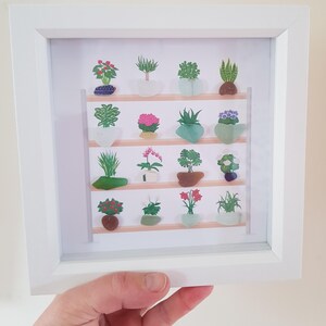 May include: Framed artwork showing miniature potted plants on wooden shelves. The plants are in various colours and shapes, within a white frame. The artwork is held by a person. The artwork is a decorative piece.